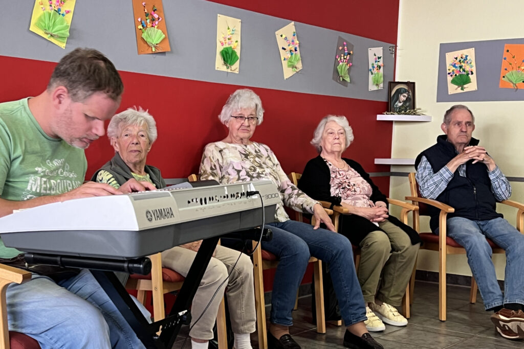 Ein Mann spielt auf dem Keyboard. Eine ältere Frau schaut ihn an. Drei andere ältere Menschen sitzen dabei.